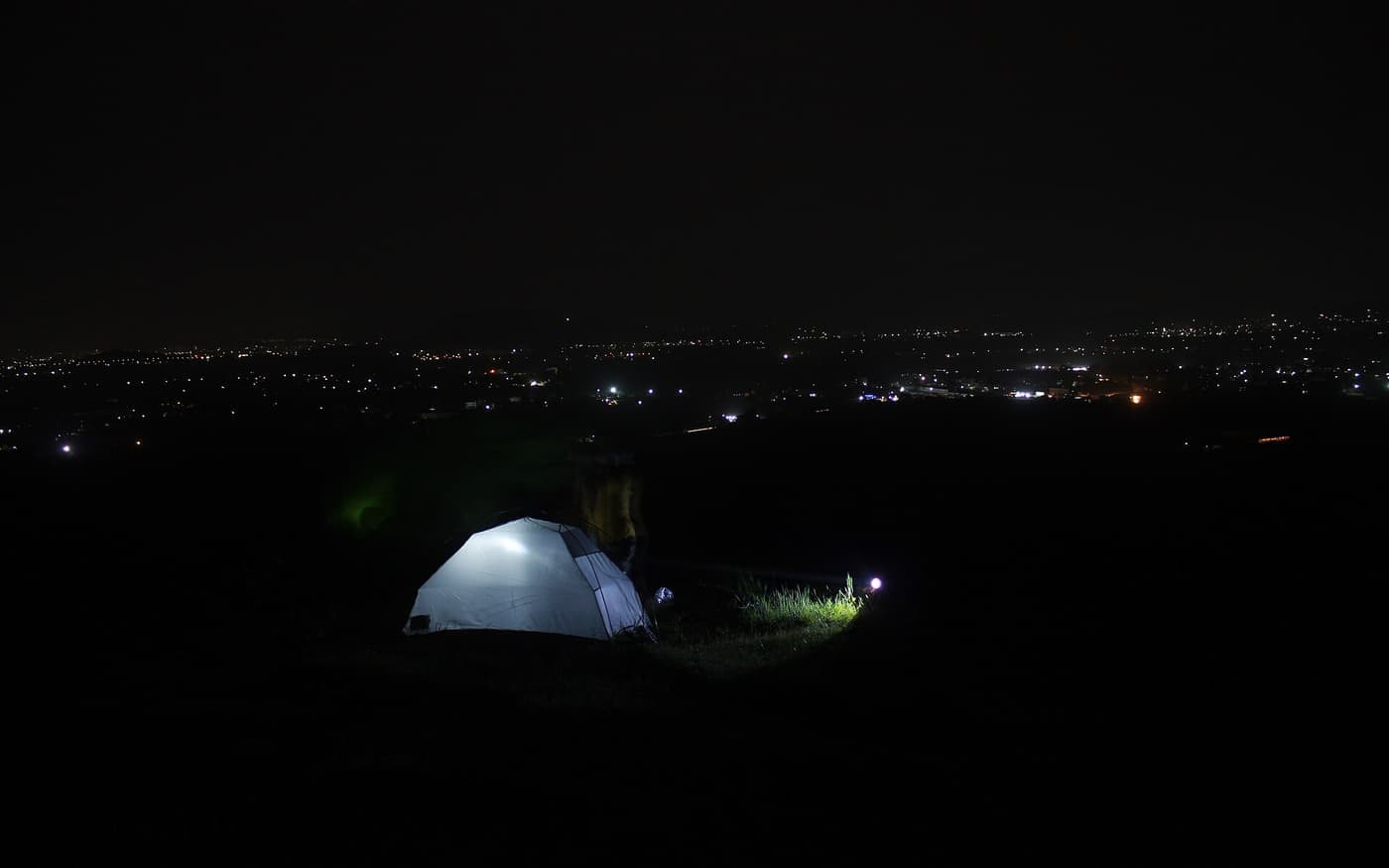 A lit tent pitched on a hillside above town lights at night