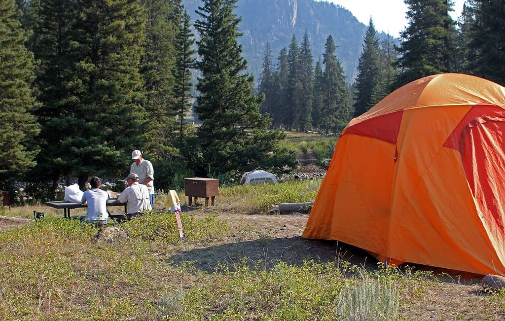 An orange family dome tent pitched at a wooded campground site, with campers sitting at a picnic table nearby